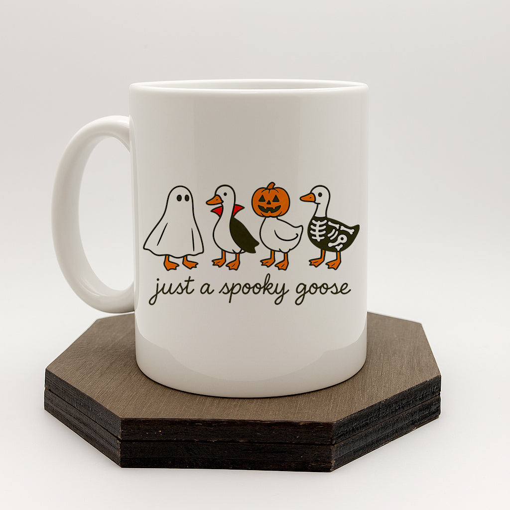 A white ceramic mug with a Halloween-themed design featuring a skeleton goose, a jack-o-lantern, and a ghost. The mug has the text "just a spooky goose" printed on it.