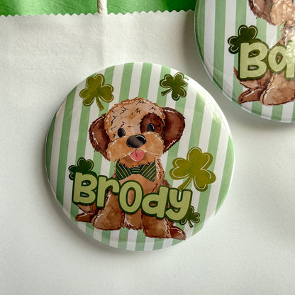 A round pin with a cartoon image of a dog wearing a green bow tie and surrounded by shamrocks.