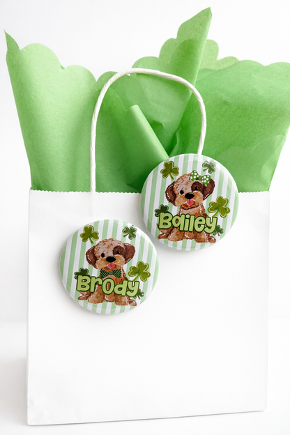 A white gift bag with a green ribbon, containing two round stickers featuring a cartoon dog and the name "Brody" written on them.