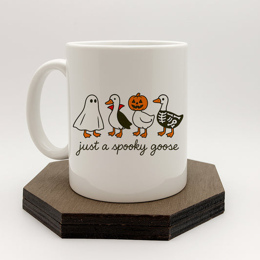 A white ceramic mug with a Halloween-themed design featuring a skeleton goose, a jack-o-lantern, and a ghost. The mug has the text "just a spooky goose" printed on it.