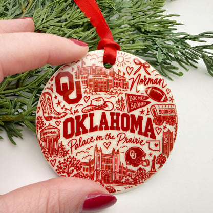 Small crimson and cream Oklahoma Sooners ceramic ornament held between fingers to show scale, featuring OU, Boomer Sooner, Norman, and campus illustrations.