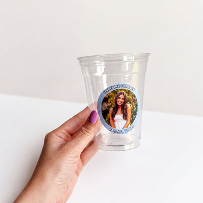 A hand holding a clear plastic cup with a sticker featuring a photo of a woman.