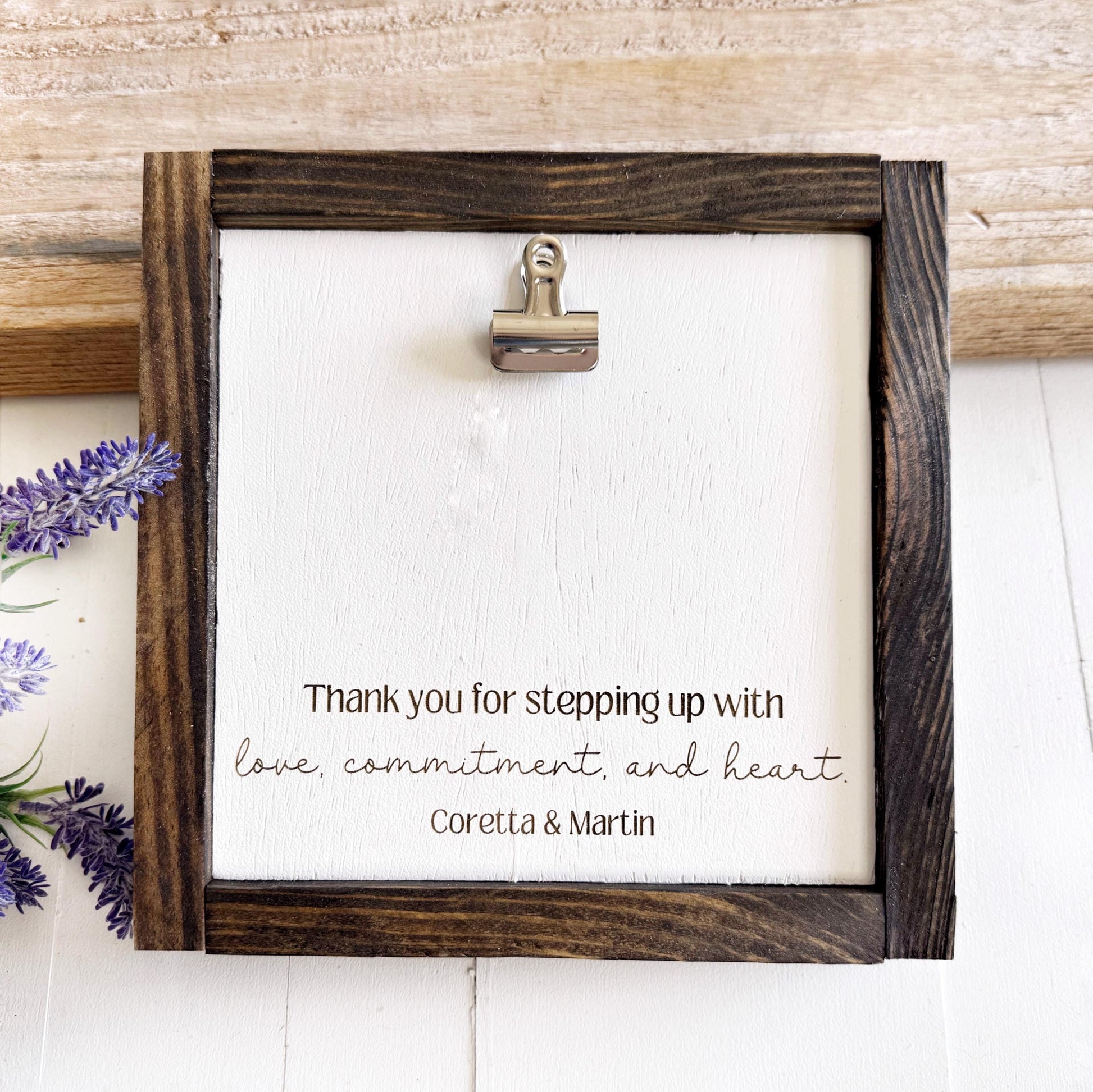 a wooden frame with a white background, and there is a silver clip attached to the frame. The frame has a message written on it, expressing gratitude for someone's commitment and love.