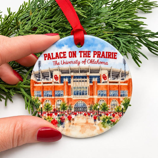 Small round ceramic OU Christmas ornament held between fingers, featuring watercolor artwork of Gaylord Family Oklahoma Memorial Stadium and Palace on the Prairie lettering.
