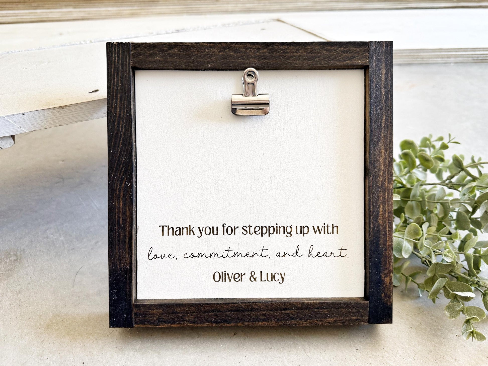 a wooden frame with a white canvas inside, displaying a heartfelt message that reads "Thank you for stepping up with love, commitment, and heart. Oliver & Lucy".