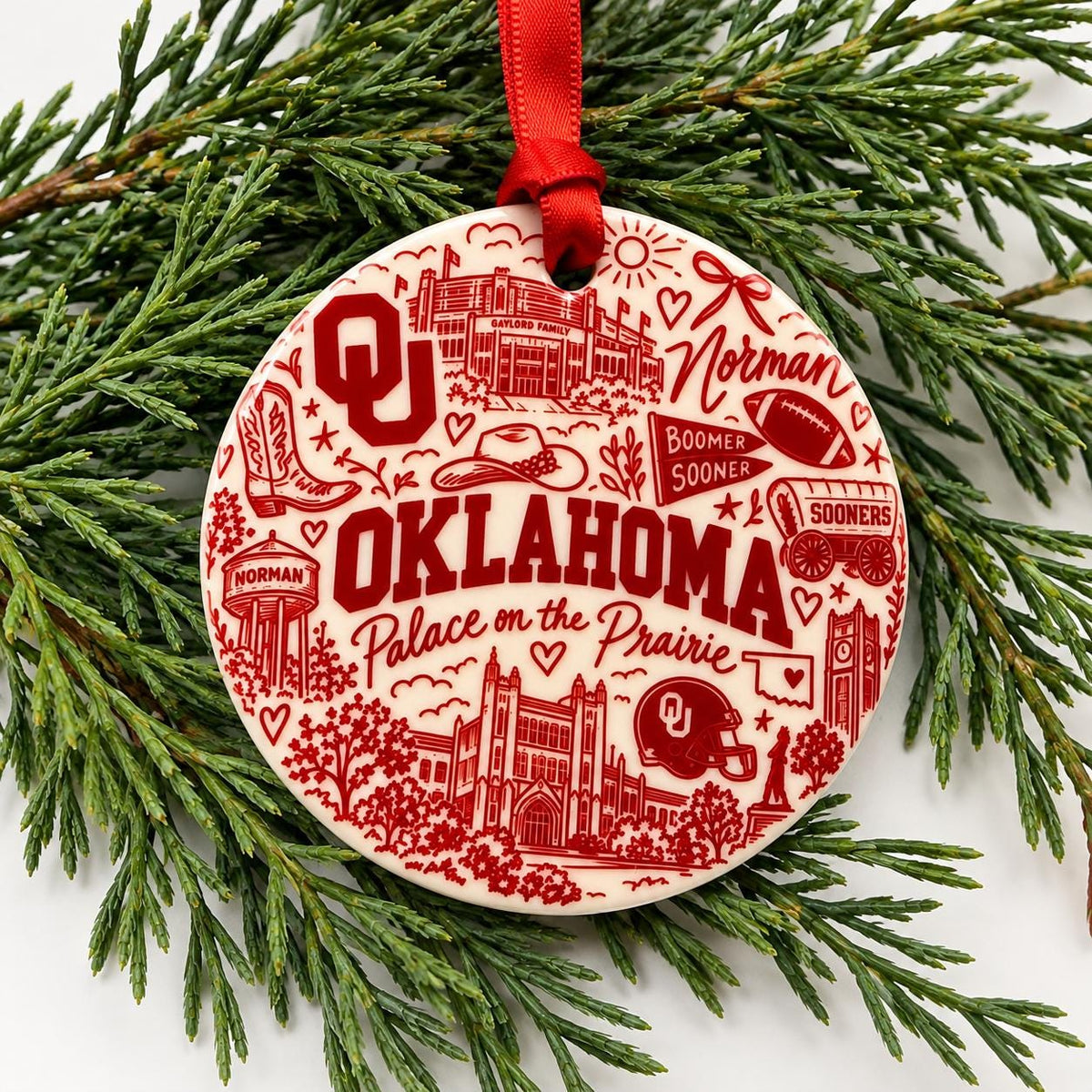 Round University of Oklahoma ceramic Christmas ornament with crimson Sooner-themed collage artwork and red hanging ribbon displayed on evergreen branches.