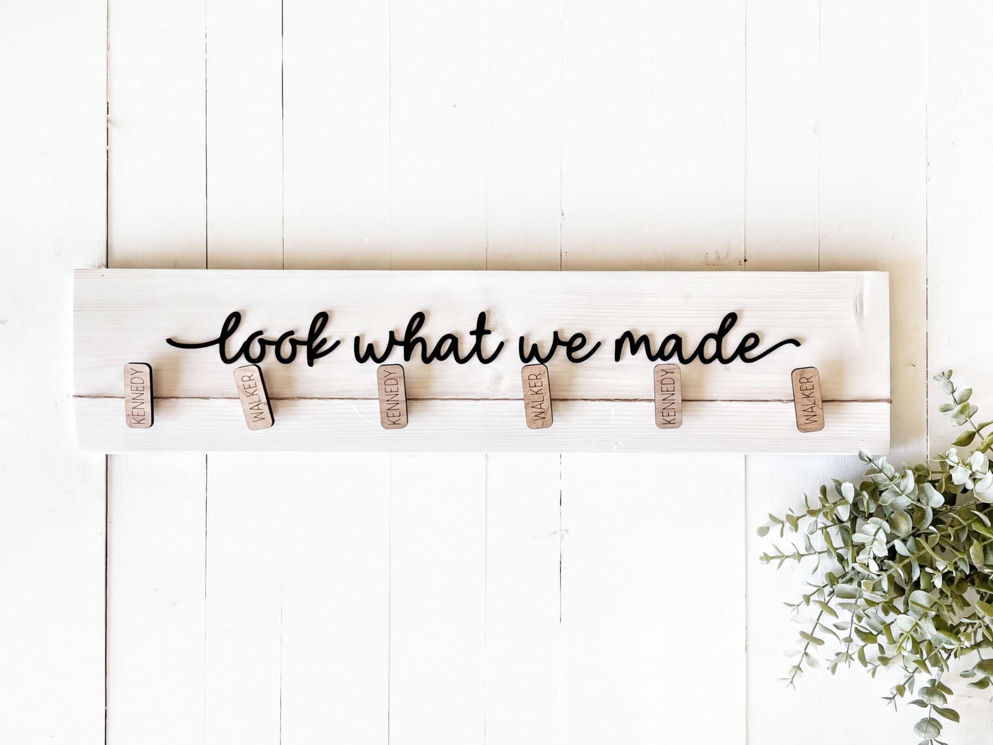 A wooden sign with the phrase "look what we made" written on it, with wooden clothespins attached to it. There is also a plant or greenery visible in the bottom right corner of the image.