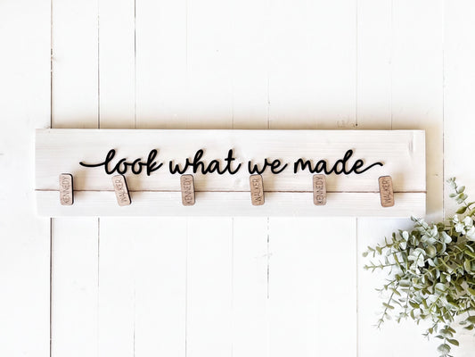 A wooden sign with the phrase "look what we made" written on it, with wooden clothespins attached to it. There is also a plant or greenery visible in the bottom right corner of the image.