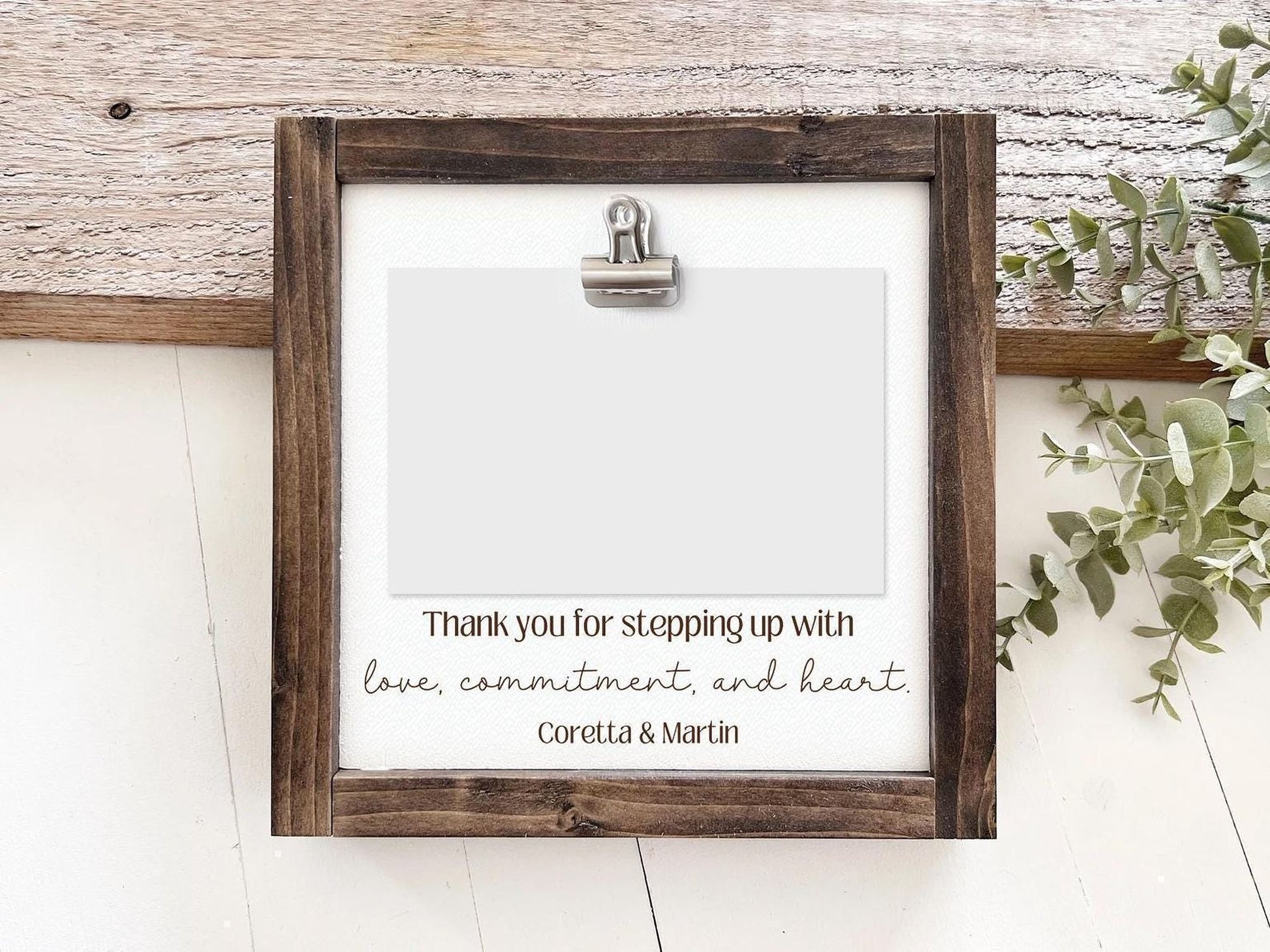 a wooden frame with a blank white card inside, placed on a wooden surface. The frame has a metal clip on the top, and there is a handwritten message on the card that reads "Thank you for stepping up with love, commitment, and heart. Coretta & Martin".