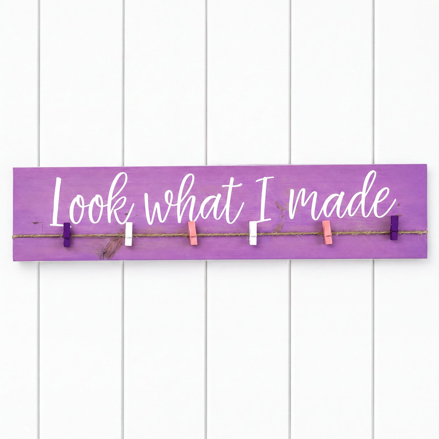 a purple sign with the phrase "Look what I made" written on it, attached to a white wall with clothespins.