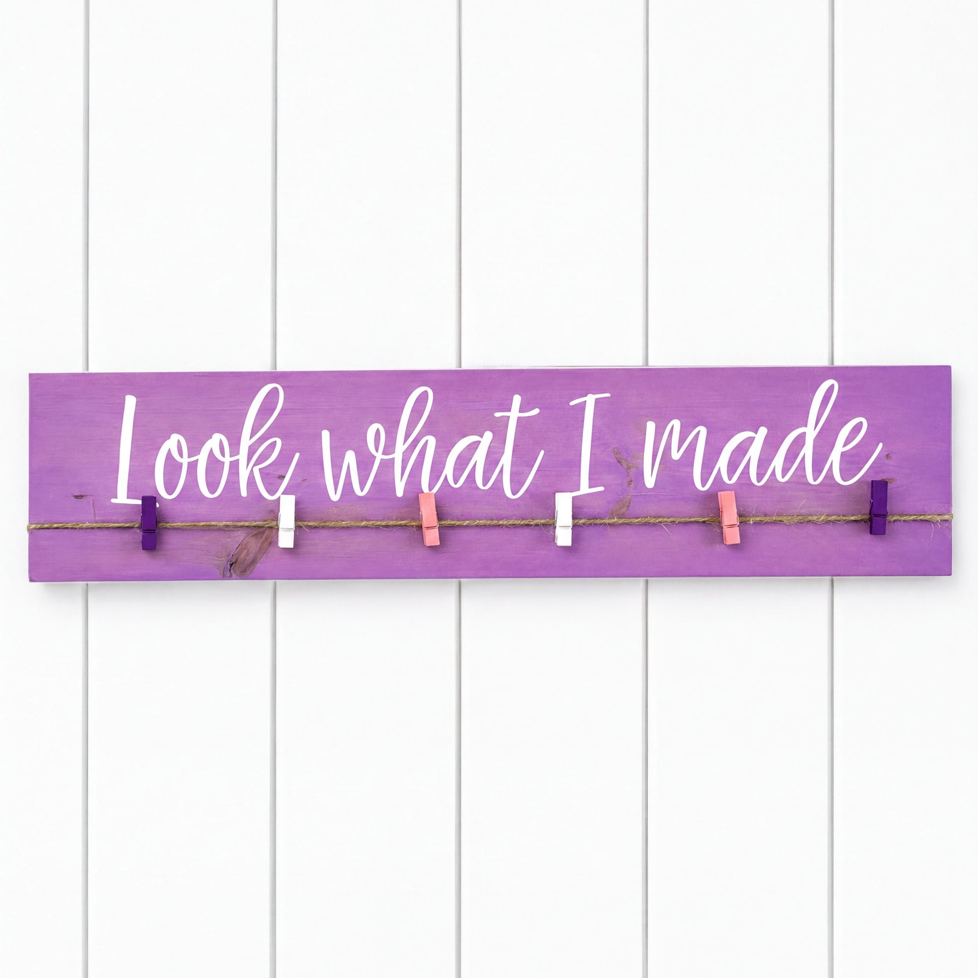 a purple sign with the phrase "Look what I made" written on it, attached to a white wall with clothespins.
