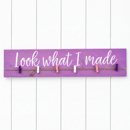 a purple sign with the phrase "Look what I made" written on it, attached to a white wall with clothespins.