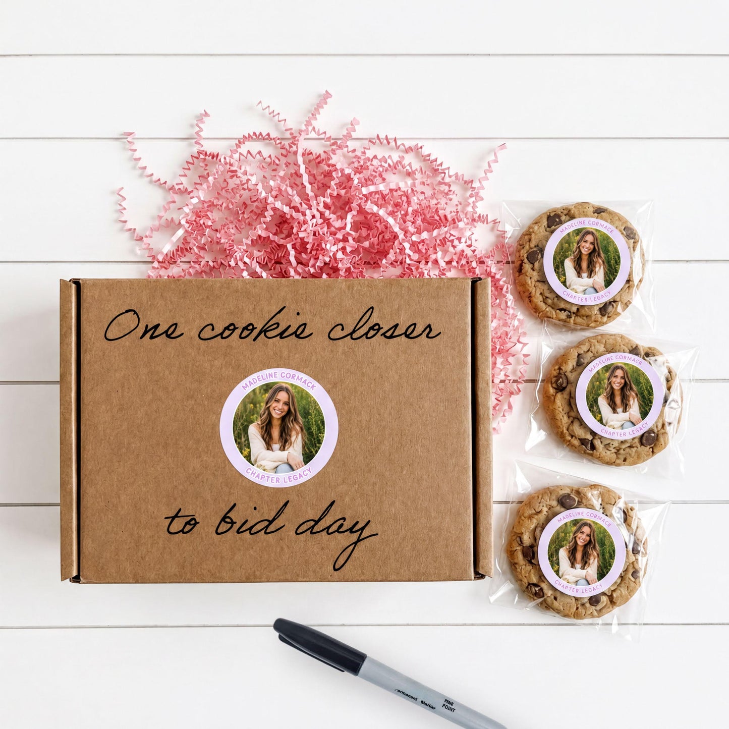 a brown cardboard box with a handwritten message, a pen, and three chocolate chip cookies with a sticker.