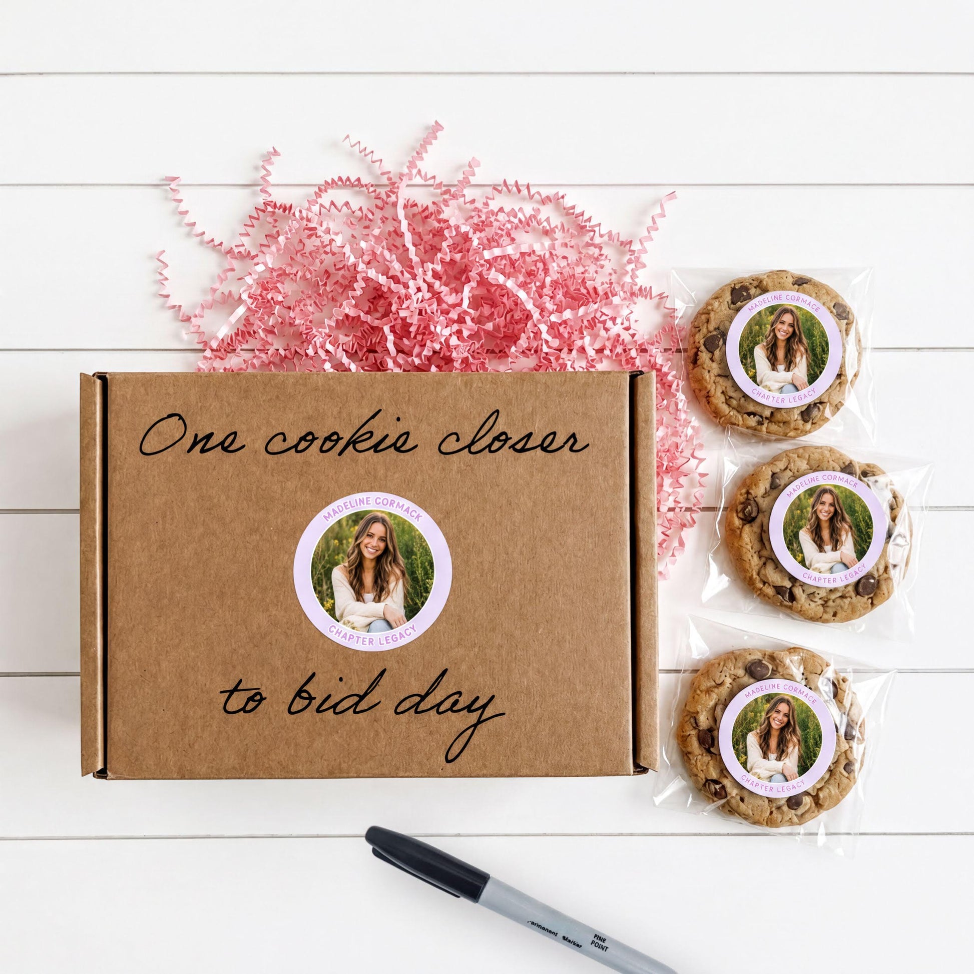 a brown cardboard box with a handwritten message, a pen, and three chocolate chip cookies with a sticker.