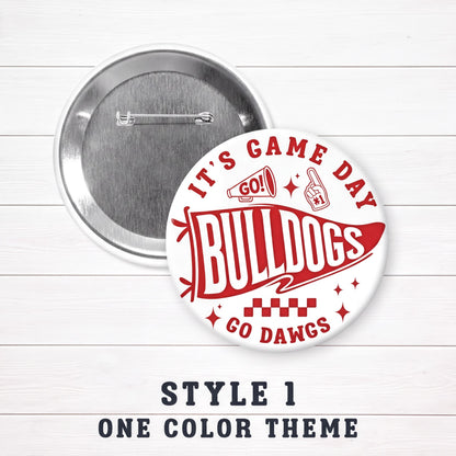 A round pin with a red and white design featuring the text "IT'S GAME DAY BULLDOGS GO DAWGS" and a football fan icon. The pin is set against a white background, and there is a caption below it that reads "STYLE 1 ONE COLOR THEMES".
