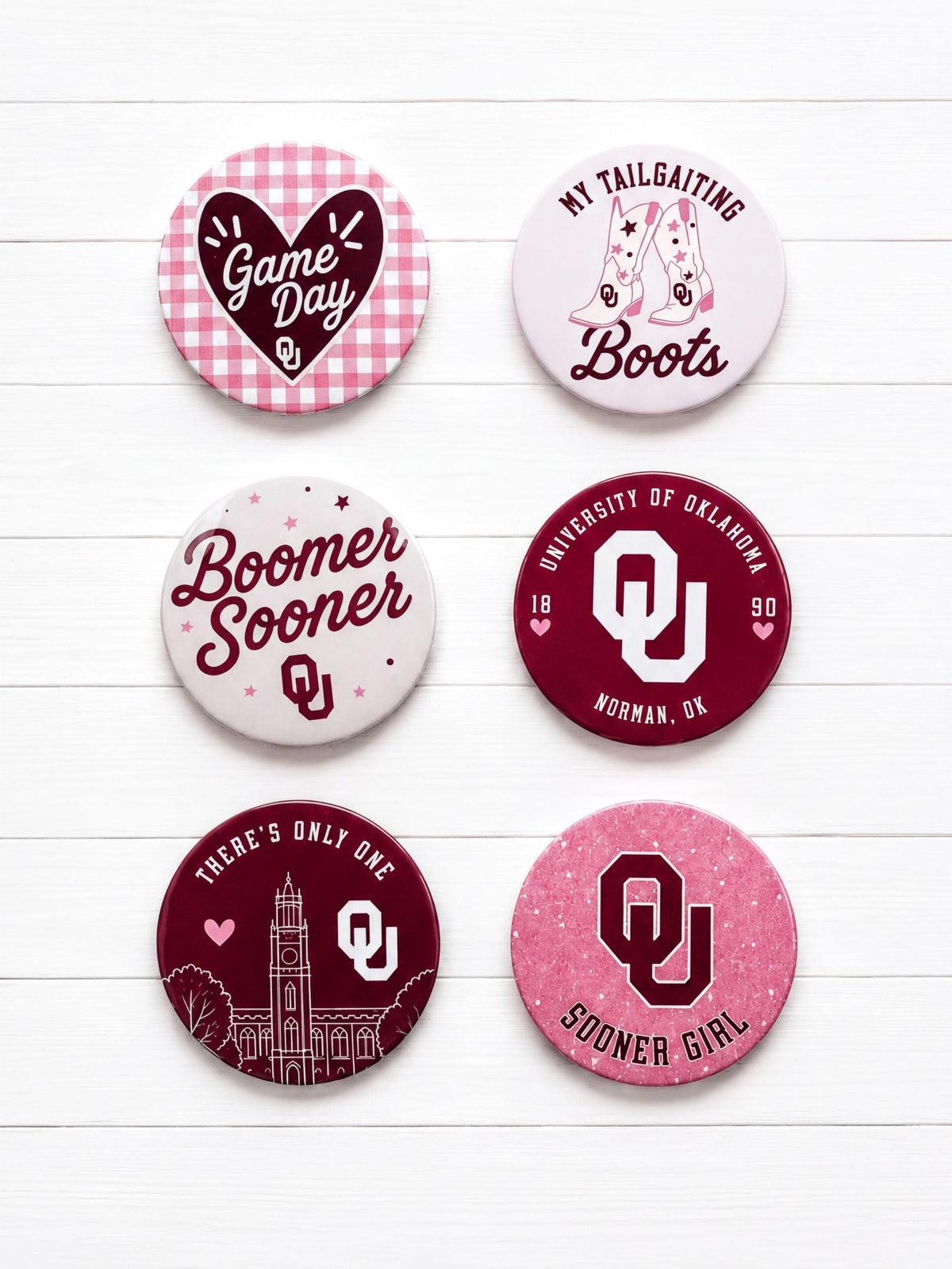 Set of OU Sooners pinback buttons featuring Boomer Sooner, Sooner Girl, tailgating boots, gingham gameday heart, University of Oklahoma seal, and campus illustration designs in crimson, white, and pink for OU gameday and spirit wear.