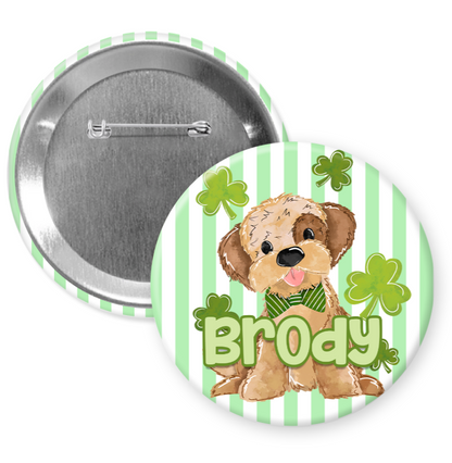 A round pin with a cartoon image of a dog wearing a green bow tie and surrounded by shamrocks. The pin has the name "Brody" written on it.