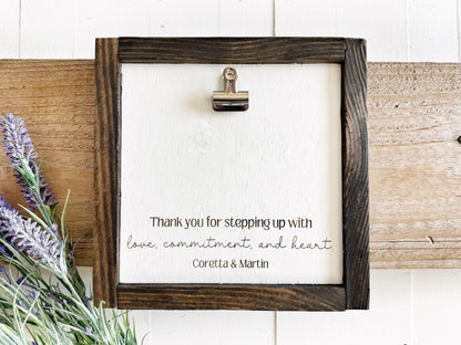 a wooden frame with a white background, and there is a silver clip on the top left corner. The frame has a handwritten message that reads, "Thank you for stepping up with love, commitment, and heart. Coretta & Martin".