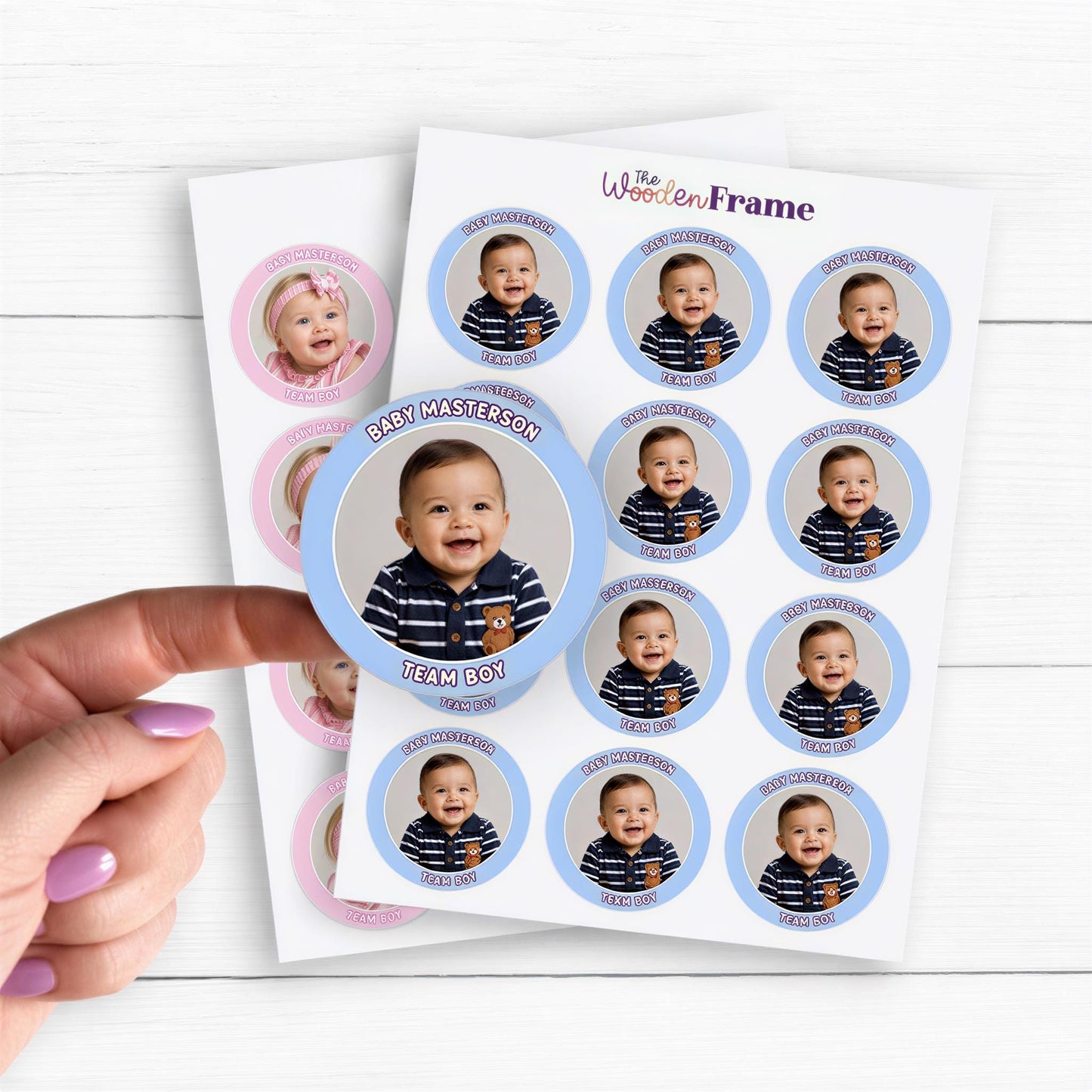 Two sheets of custom gender reveal stickers featuring baby boy and baby girl photos on round glossy labels. Blue “Team Boy” stickers show a smiling baby with a navy striped shirt and “Baby Masterson” text. Pink “Team Girl” stickers show a baby girl in a pink outfit. A hand lifts one sticker on a white wooden background, showcasing personalized photo stickers for gender reveal parties, baby showers, favors, and decorations.