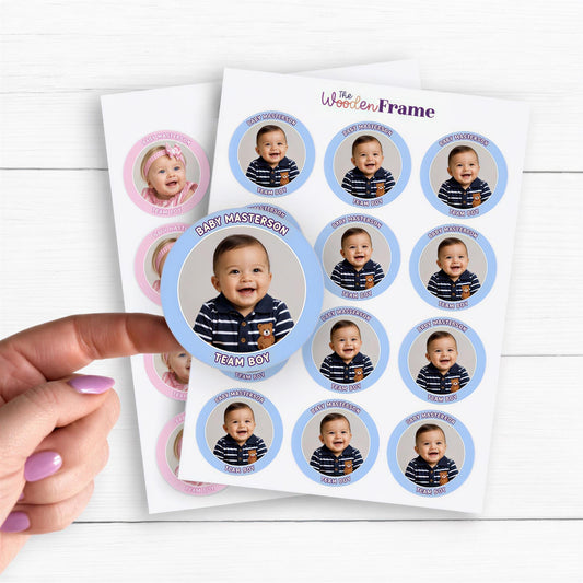 Two sheets of custom gender reveal stickers featuring baby boy and baby girl photos on round glossy labels. Blue “Team Boy” stickers show a smiling baby with a navy striped shirt and “Baby Masterson” text. Pink “Team Girl” stickers show a baby girl in a pink outfit. A hand lifts one sticker on a white wooden background, showcasing personalized photo stickers for gender reveal parties, baby showers, favors, and decorations.